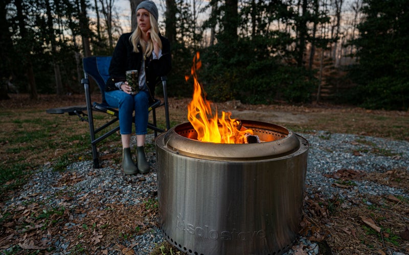 A woman sitting behind a fire pit with trees in the background