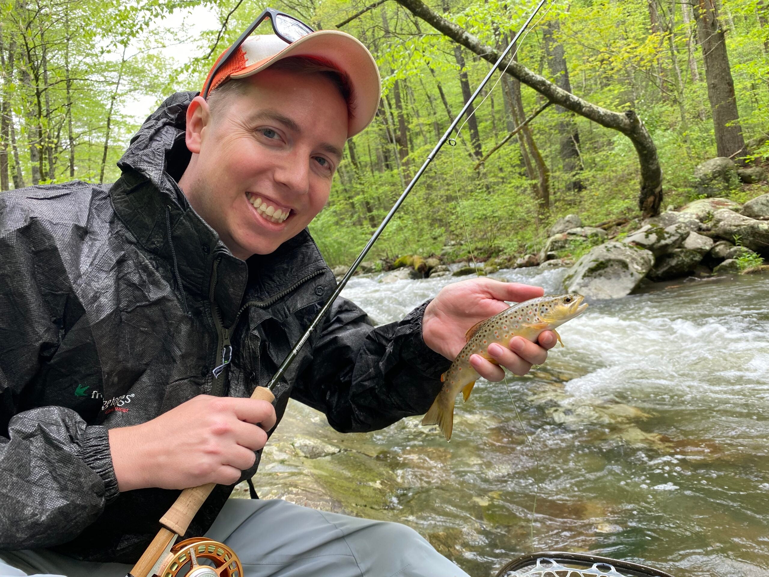 Man holding small trout next to a rushing stream in summer