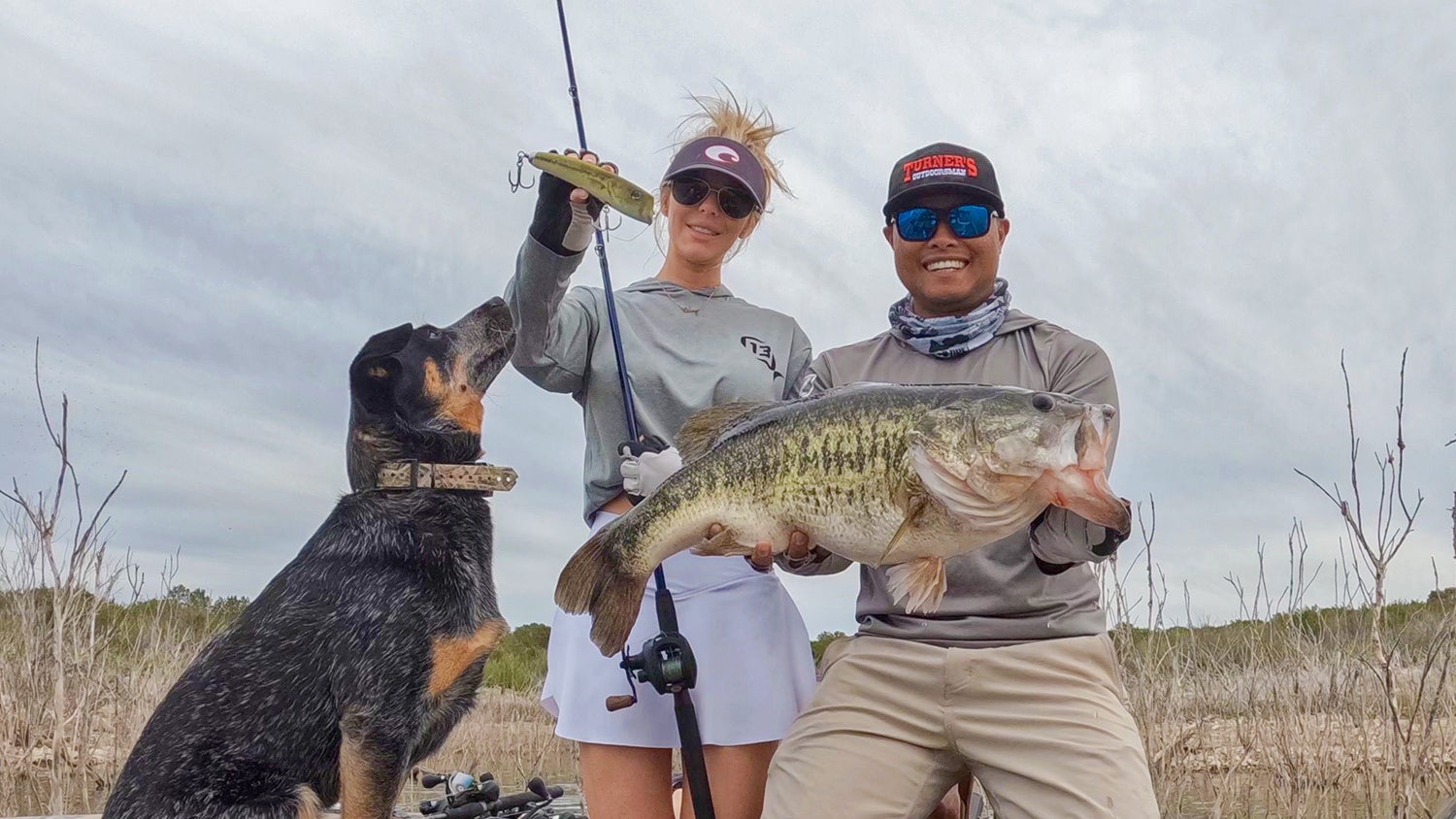 two anglers, one with huge largemouth, the second holding a large lure. sitting dog stares at lure.
