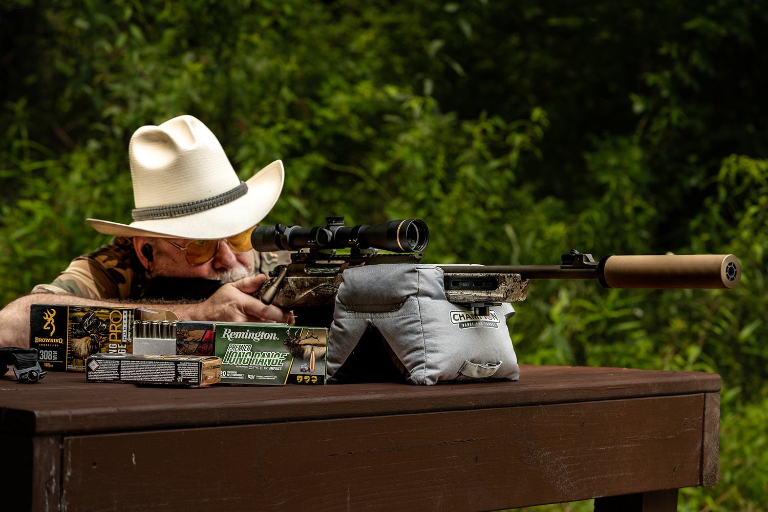 shooter uses bench and sandbags for support as he aims rifle with suppressor; ammunition sits in front of gun