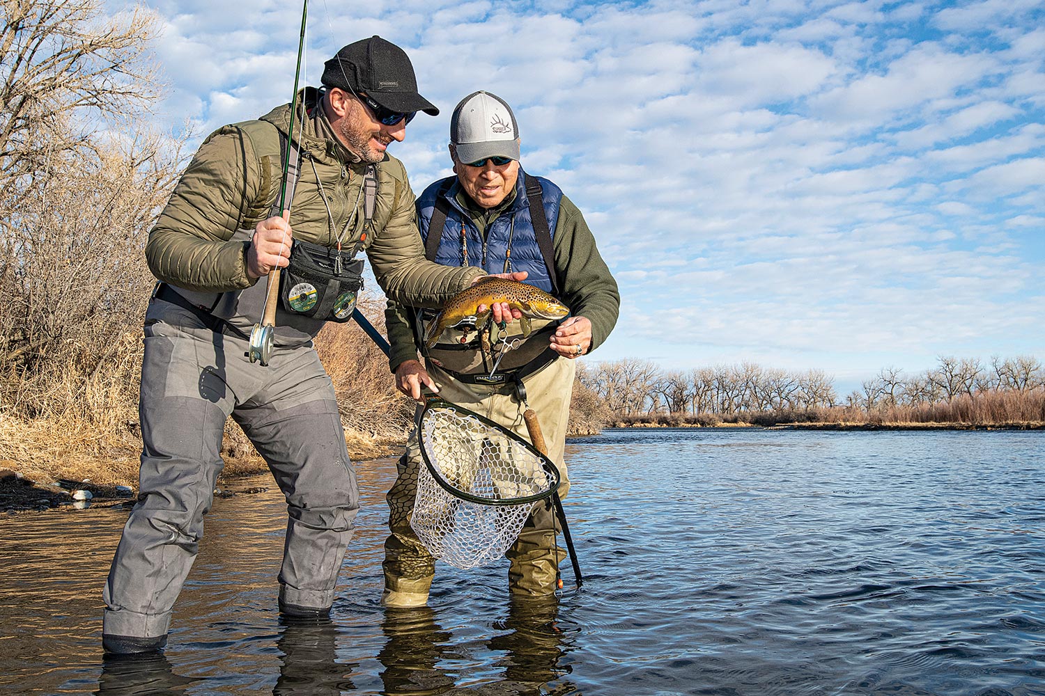 standing in river near bank, one angler holds a brown trout, another holds a net