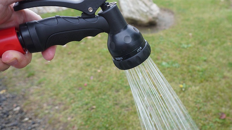 The red and black nozzle of the Klymit WaterPod shower system spraying water in front of a green grass background.