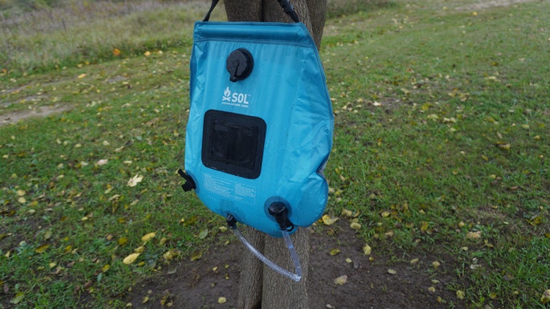A blue and white SOL solar shower hanging from a tree against a green grass background.